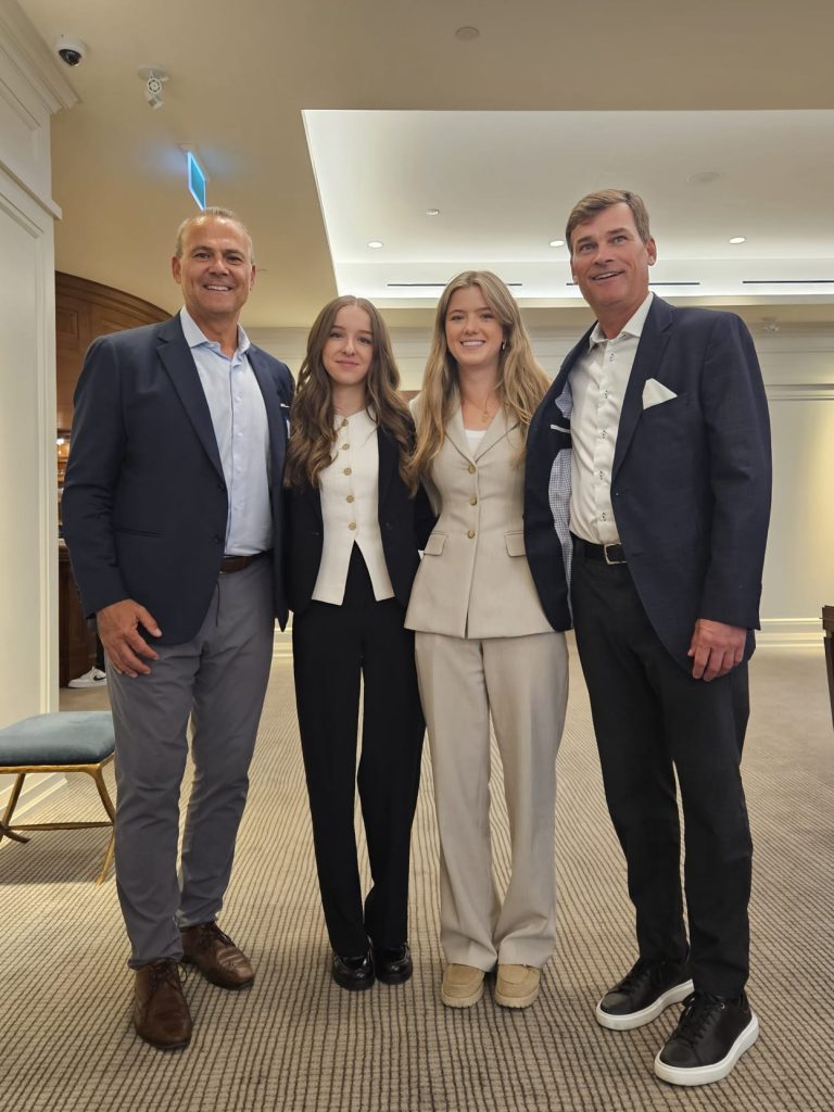 Two Barometer Capital interns stand between two senior team members, all dressed in professional business attire, smiling for a group photo at the Invested & Connected event. The setting is a well-lit conference space with neutral-toned decor, highlighting a moment of mentorship and collaboration across generations.